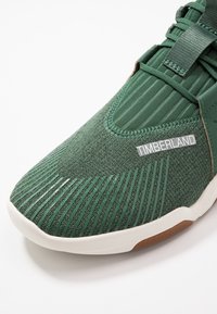 Baskets textiles vertes avec texture nervurée, semelle blanche et logo Timberland. Arborent un design épuré et un laçage élastique pour un maintien sécurisé.