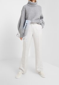 Harmaa paksu neulottu pooloneule, valkoiset räätälöidyt housut, kädessä kirkas laukku, yhdistetty valkoisiin tennareihin vaalealla taustalla.