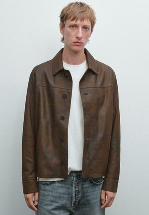 Young person with short blonde hair wearing a brown button-up leather jacket over a white shirt and faded blue jeans, standing against a plain background.
