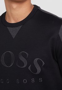 Svart sweatshirt med en texturerad yta, med en kontrasterande mörkgrå V-formad detalj vid kragen och broderad logotyp framtill.