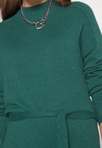 Maglione lavorato a maglia color teal con scollatura a coste, maniche lunghe, lacci in vita e una collana con ciondolo a forma di cuore metallico.