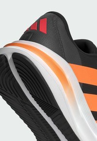 Zwarte sportieve schoen met een oranje en witte tussenzool, voorzien van gestructureerde stof, rode accenten en een opvallende zwarte rubberen loopzool met groeven.