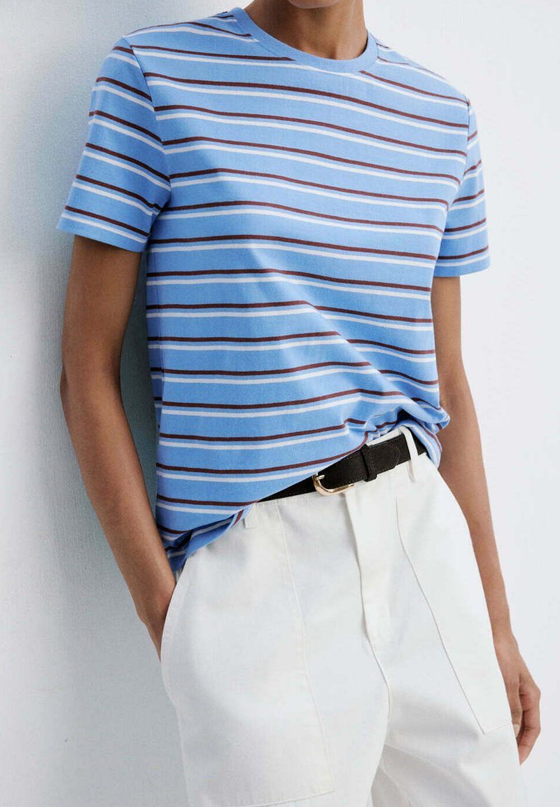 Personne portant une chemise à rayures bleues à manches courtes rentrée dans un pantalon blanc avec une ceinture noire, debout avec une main dans la poche.