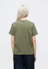 Olīvu zaļš kokvilnas T-krekls ar īsām piedurknēm, apaļu kakla izgriezumu un brīvu piegriezumu, redzams no aizmugures, saskaņots ar tumšiem džinsiem.