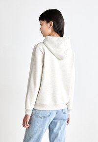 Uma hoodie bege claro feita de tecido suave, com capuz ajustável, punhos e barra canelados. Usada com jeans claros desgastados.