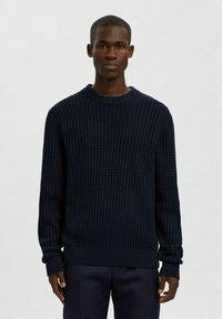 Uomo che indossa un maglione a maglia testurizzata blu navy scuro e pantaloni abbinati, in piedi davanti a uno sfondo chiaro e neutro.