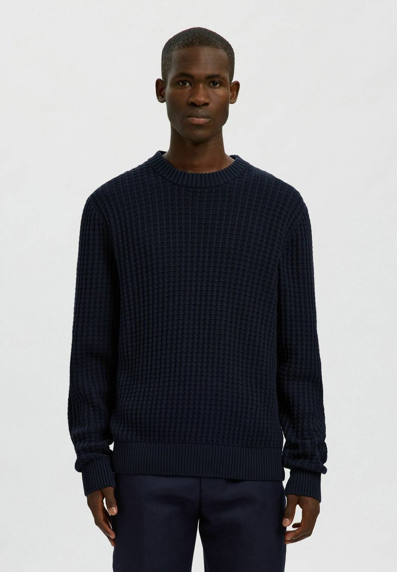Uomo che indossa un maglione a maglia testurizzata blu navy scuro e pantaloni abbinati, in piedi davanti a uno sfondo chiaro e neutro.
