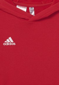 Sweatshirt à capuche rouge en tissu doux, avec un logo Adidas brodé en blanc sur la poitrine gauche. Texture lisse avec un design simple.