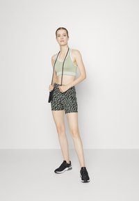 Sutiã desportivo verde claro com detalhes brancos, emparelhado com calções pretos com padrão. Modelo usa sapatilhas de atletismo pretas e segura uma corda de saltar.