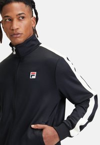 Zwarte zip-up trainingsjack met witte zijstippen, voorzien van een borstlogo en stoffen zakken, gladde textuur en een getailleerd design.