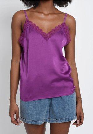 Mujer vestida con una camiseta de satén púrpura con ribete de encaje y shorts de mezclilla azul, de pie contra un fondo blanco liso.