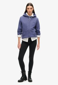 Sudadera con cremallera morada, con cordón y logotipo bordado, combinada con una camisa blanca de cuello, jeans ajustados negros y botas negras de cordones.