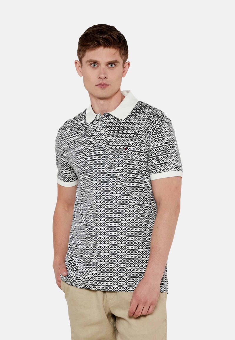 Camisa polo de patrón en blanco y negro con cuello y mangas cortas. Presenta un ribete blanco y un pequeño logo en el pecho.
