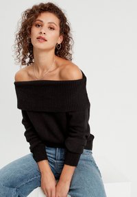 Sort off-shoulder sweater med ribbet halsudskæring og manchetter, parret med lyseblå jeans. Har et blødt, tekstureret strikstof.