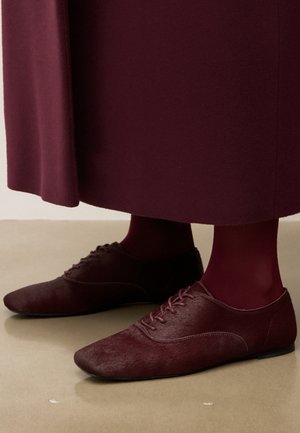 Burgundin väriset nauhakengät, jotka on yhdistetty samansävyisiin sukkahousuihin ja pitkään burgundinväriseen takkiin, seisoo sileällä beigeellä lattialla.