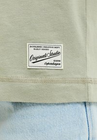 Μπεζ τελείωμα πουκάμισου με ραμμένη λευκή ετικέτα που γράφει "Jack & Jones Creative Dept., Originals Studio, Κοπεγχάγη, 1990" πάνω σε ανοιχτόχρωμο μπλε τζιν.