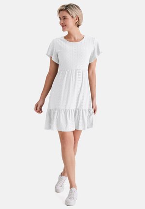 Frau mit kurzen blonden Haaren, die ein weißes Kleid mit Lochmuster und weiße Turnschuhe trägt, geht und schaut seitlich vor einem schlichten Hintergrund.