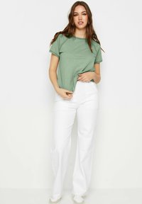 T-shirt verde di cotone con maniche corte e cuciture decorative sulle spalle, abbinato a pantaloni bianchi a vita alta e sneakers.