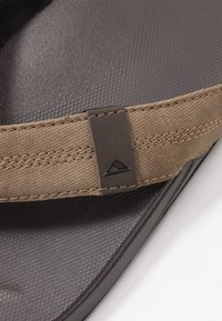 Tongs marron avec une semelle en caoutchouc texturée. Elles présentent une sangle en tissu beige avec des détails en surpiqûre et un petit logo noir.