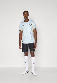 Ανοιχτό μπλε αθλητική φανέλα με σχέδιο και κοντά μανίκια, μπεζ λεπτομέρειες, μαύρο λογότυπο, φορεμένη με μαύρες βερμούδες και λευκές κάλτσες, συνδυασμένη με παπούτσια ποδοσφαίρου.