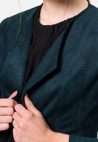 Turquoise suède jas met een gestructureerd ontwerp en opvallende naden, voorzien van een reverskraag en gedragen over een zwarte top.