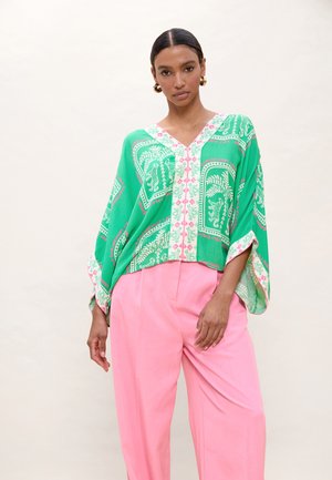 Mujer con blusa suelta de cuello en V con estampado verde, mangas anchas y pantalones rosas de cintura alta, de pie frente a un fondo liso y claro.