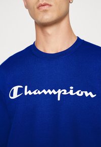 Královsky modrá mikina s bílým logem "Champion" v kurzivním písmu. Tkanina vypadá jemně a má klasický design s kulatým výstřihem.