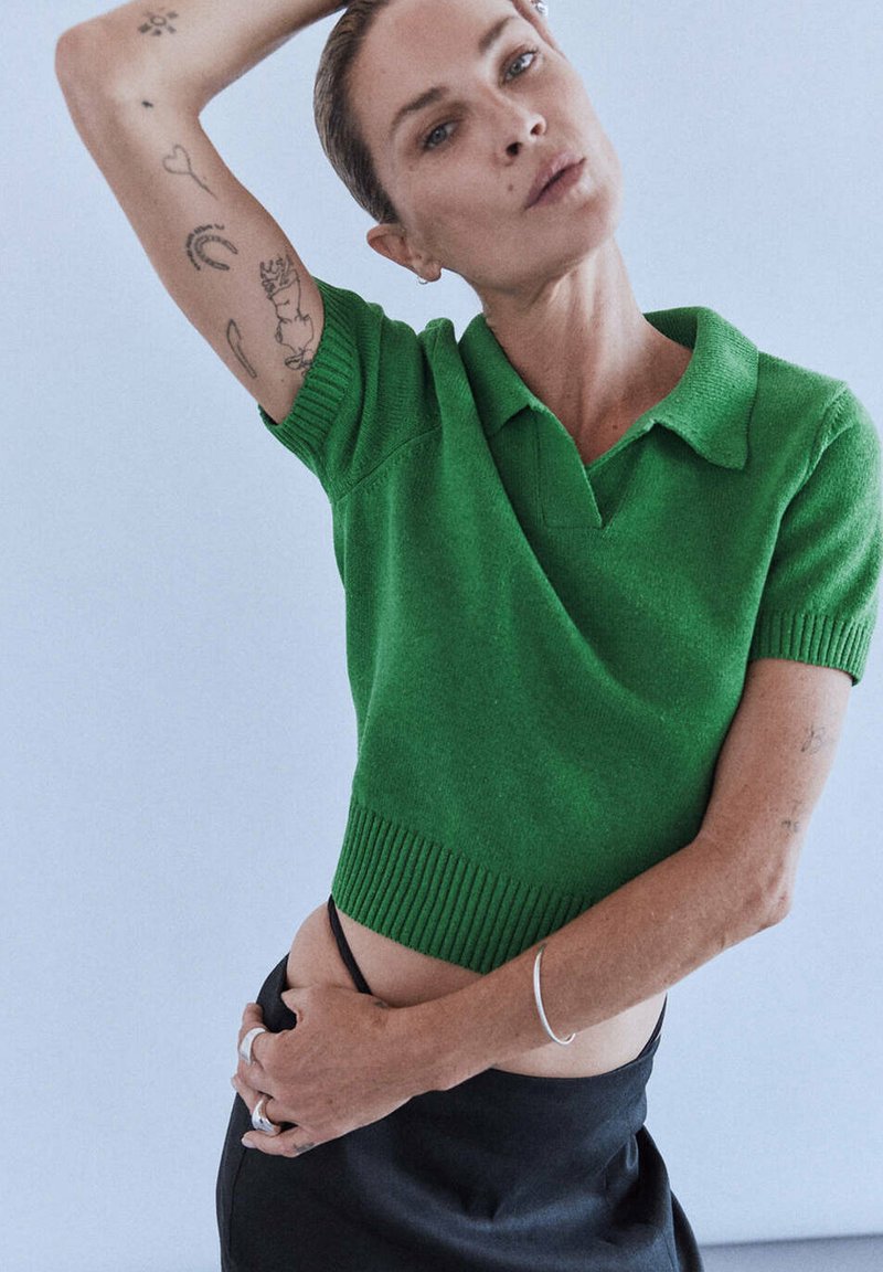 Personne portant un pull vert à manches courtes et une jupe noire, levant un bras montrant des tatouages sur son avant-bras sur un fond clair.