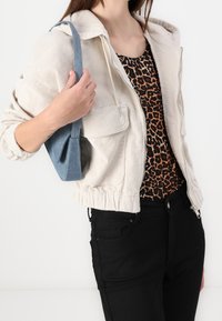 Kvinna som bär en beige kordjacka över en leopardmönstrad topp, svarta byxor och håller en liten blå denimaxelväska på sin axel.