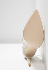 Nude patenterad läder stiletto-klack med spetsig tå, med en slät yta och minimalistisk design, lutad mot en vägg.