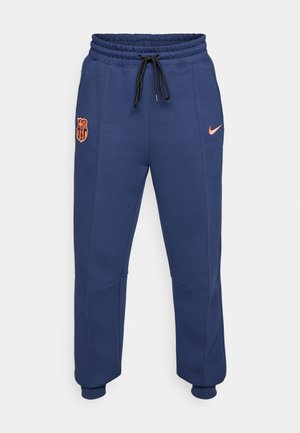 Calças de fato de treino azul-marinho feitas de material macio, com um cós elástico e cordão, bolsos laterais e um logo laranja da Nike com um emblema do Barcelona.