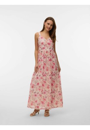 Kvinne som smiler, står og holder forsiktig en rosa blomstret ermeløs maxikjole, iført beige sandaletter med hæl.