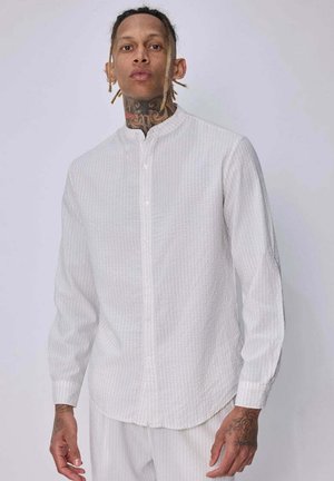 Homme avec des tatouages portant une chemise blanche à manches longues boutonnée et un pantalon blanc assorti sur un fond clair uni.