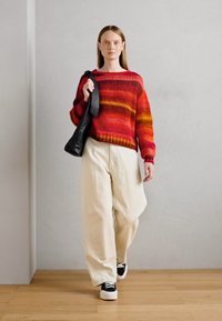 Gebreide trui met rode en oranje strepen, gecombineerd met beige wijde broek en zwarte sneakers. Draagt een zwarte schoudertas.