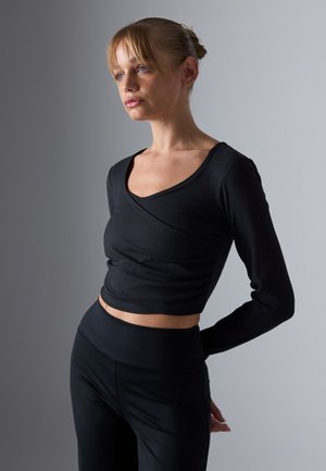 Jongeman met blond haar in een knot, draagt een op maat gemaakte zwarte lange mouwen crop top en zwarte high-waisted broek, staand met de handen achter de rug.