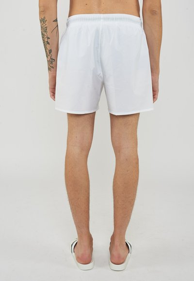 Homme portant un short blanc et des sandales blanches, debout dos à la caméra, montrant un tatouage sur le bras gauche.
