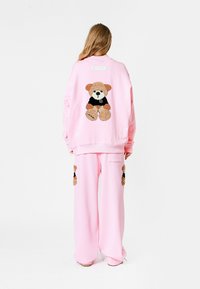 Conjunto de sudadera y pantalones de gran tamaño en color rosa, con un aplique de oso de peluche en la parte posterior y bolsillos laterales con detalles similares de oso de peluche.