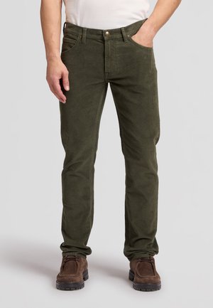 Homme portant un pantalon en velours côtelé vert olive et des chaussures marron à lacets, une main dans la poche et une chemise blanche rentrée.
