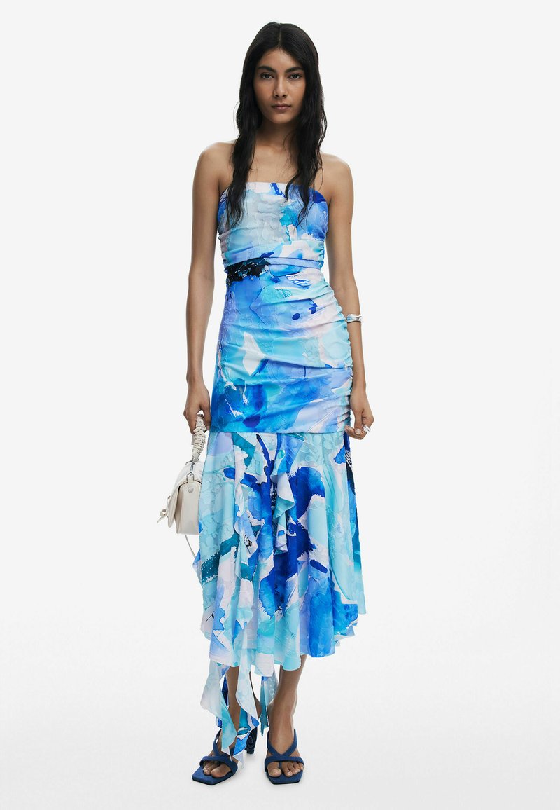 Robe maxi à imprimé floral bleu et blanc avec un corsage ajusté, un ourlet à volants et une texture drapée ; associée à des sandales à lanières bleues et un petit sac blanc.