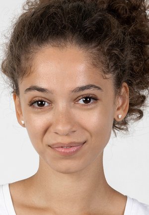 Nahaufnahme einer jungen Frau mit lockigem Haar, die ein weißes Hemd trägt. Sie hat kleine herzförmige Ohrringe und einen neutralen Hintergrund.