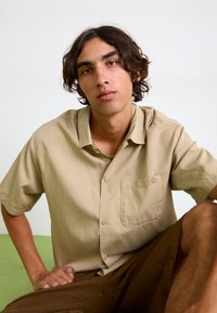 Beige short-sleeved shirt with a chest pocket, made of a smooth fabric. Accented with a small brand logo. Brown shorts, relaxed fit.
