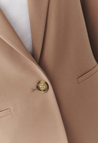 Gros plan d'un blazer beige avec un bouton en écaille de tortue et un col cranté sur un top blanc.