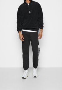 Svart fleece zip-up-tröja med ribbstickade muddar och en liten vit logotyp; kombinerad med svarta sweatpants med grafiskt tryck och vita sneakers.