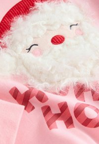 Vaaleanpunainen kangas, jossa on brodeerattu joulupukin kasvot, joissa on teksturoitu valkoinen parta, punainen hattu ja painettu "HO HO HO" makeissauvakuosisena.