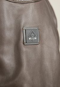 Braunes Leder mit strukturierter Oberfläche; enthält ein schwarzes Emblem mit einer diamantförmigen Gestalt und dem Gestaltungselement "1982".