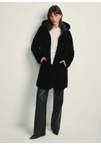Czarny futrzany płaszcz z kapturem, długimi rękawami i kieszeniami bocznymi, w połączeniu z czarnymi skórzanymi spodniami. Materiał wydaje się miękki i teksturowany.
