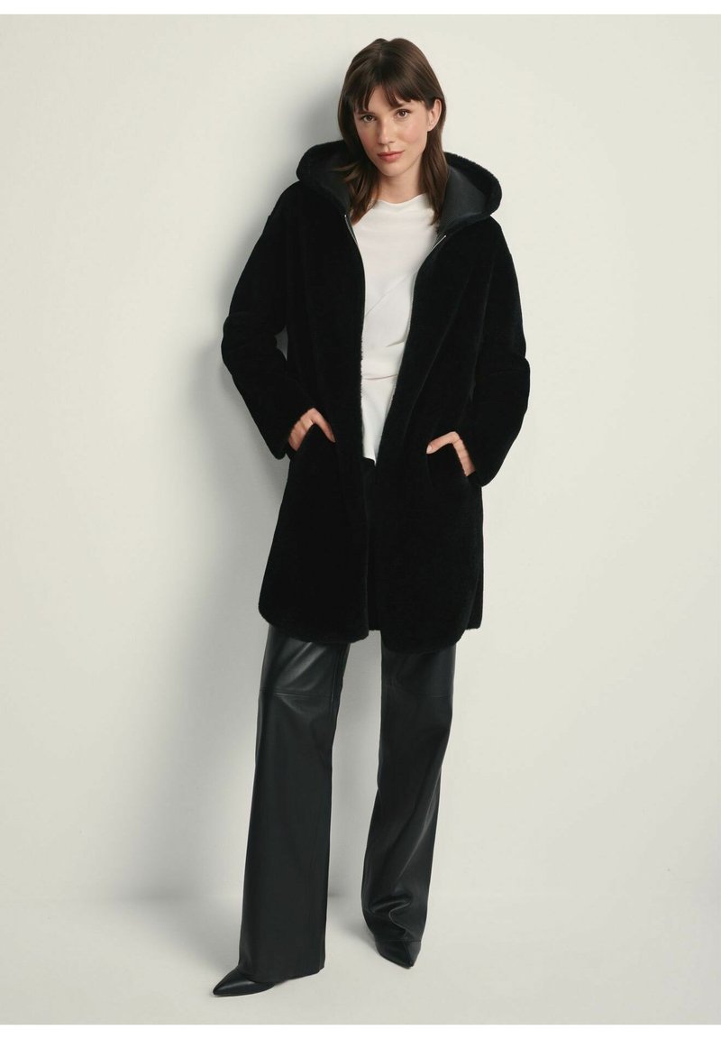Czarny futrzany płaszcz z kapturem, długimi rękawami i kieszeniami bocznymi, w połączeniu z czarnymi skórzanymi spodniami. Materiał wydaje się miękki i teksturowany.