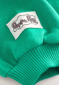 Zaļš adīts džemperis ar ribotām aprocēm, baltu etiķeti ar rozā burtiem "Peppa Pig", teksturēta auduma detaļa.