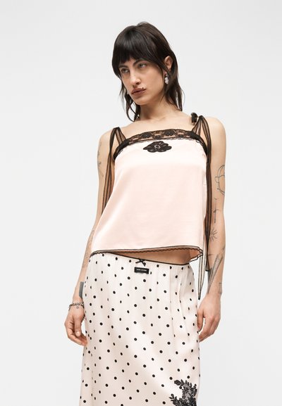 Femme portant un caraco en satin rose pâle avec bordure en dentelle noire et une jupe à pois avec broderie florale, sur fond blanc uni.