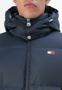 Navy puffer jacket with a high collar, featuring a front zipper, snap buttons, and a Tommy Jeans logo patch on the chest. Textured fabric.